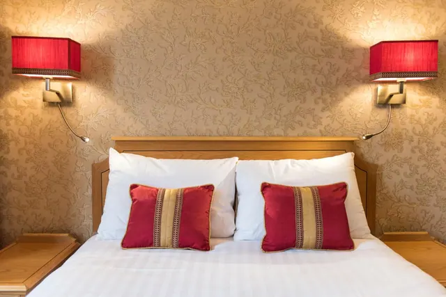 A double bed with white pillows and two red cushions, flanked by red wall lamps against patterned wallpaper.