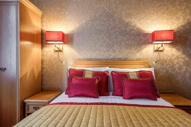 A double bed with red and gold cushions and a gold quilt, with two lit red wall lamps and a wooden wardrobe on the left.