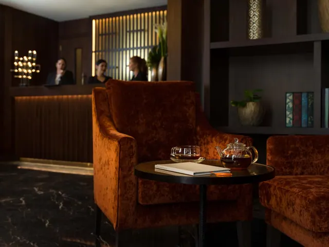 A lobby seating area with two orange velvet armchairs, a side table holding a teapot, a glass cup and a book, with reception staff blurred in the background.