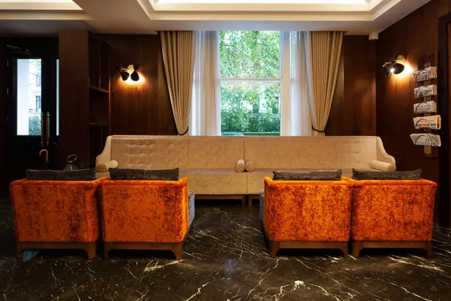 A lobby seating area with orange velvet armchairs, a beige tufted sofa, a dark marble floor and a window showing trees outside.
