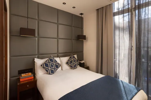A double bed with a padded grey headboard wall, two pendant lamps, and sheer curtains beside a tall window.
