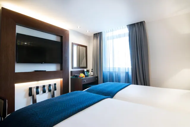 A hotel bedroom with two beds covered in blue quilted throws, a wall-mounted television, a dresser with fruit, and a window with curtains.