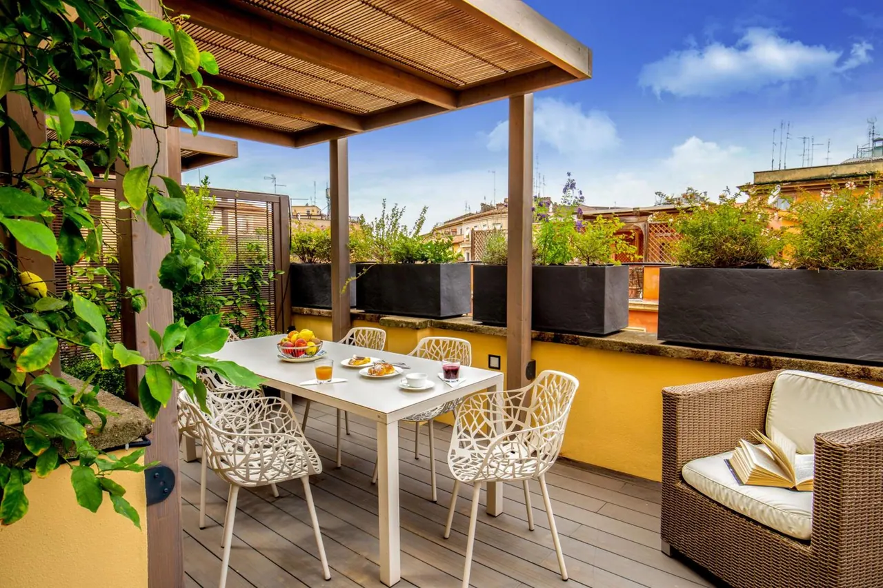 A rooftop terrace under a wooden pergola has a set breakfast table with white chairs, planters, a wicker armchair with an open book, and a view over neighbouring rooftops.