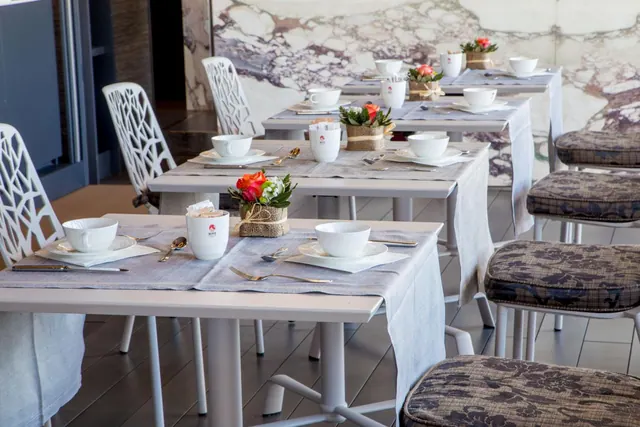 A restaurant dining room with white tables set with cups, saucers, cutlery and flower centrepieces, beside a marble wall.