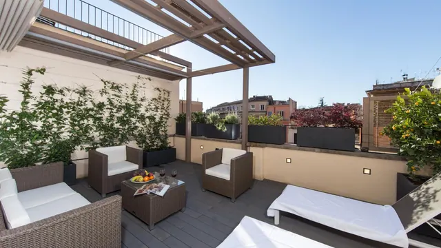 A rooftop terrace with a wooden pergola, wicker lounge chairs, sun loungers, potted plants and a view of nearby buildings.
