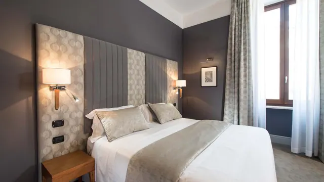 A double bed with a grey upholstered headboard, wall reading lamps, and a window with sheer curtains in a hotel bedroom.