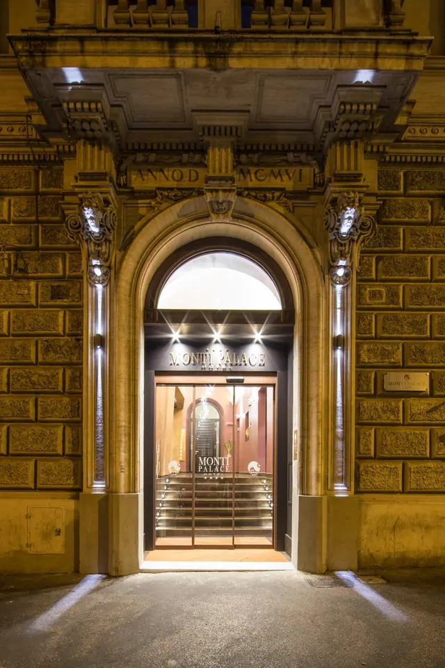 Night view of the arched stone entrance of the Monti Palace hotel, with glass doors and a lit staircase visible inside.