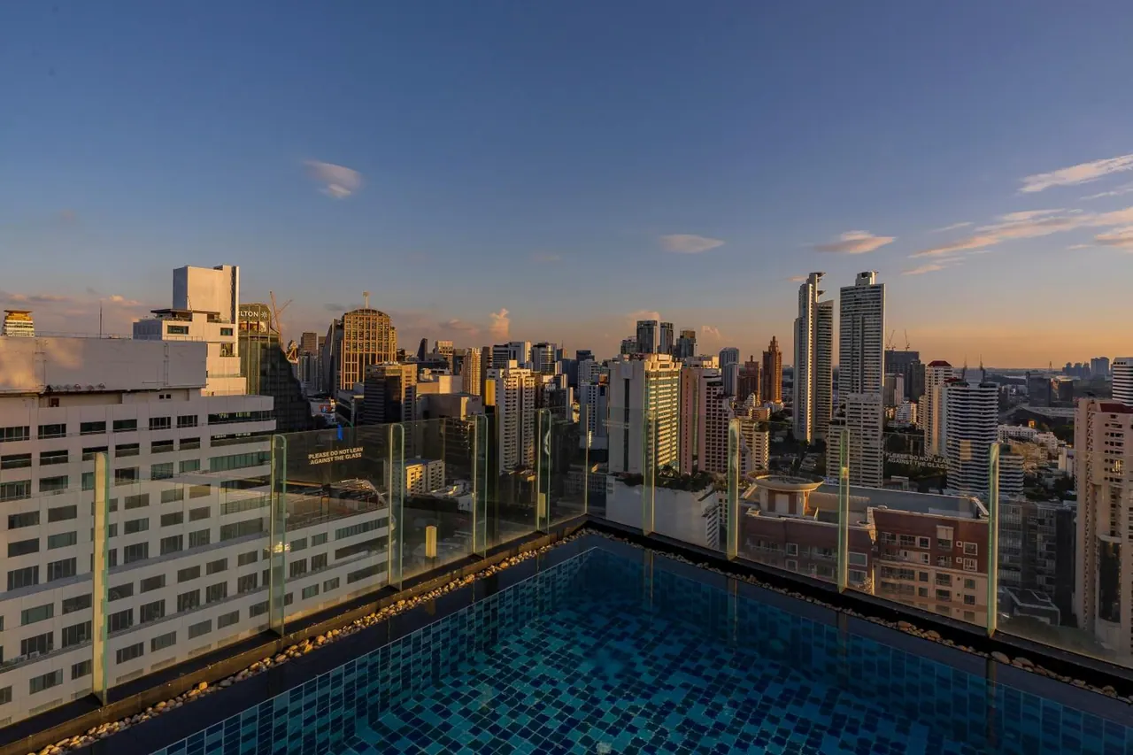A rooftop swimming pool with a glass railing overlooks a city skyline of high-rise towers at sunset.