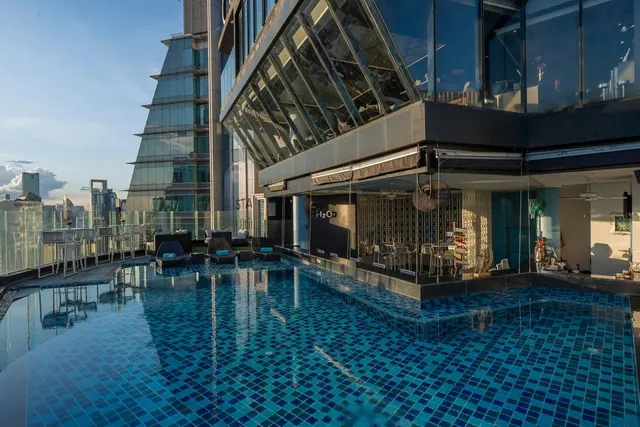 An outdoor rooftop pool with blue mosaic tiles, sun loungers and a glass-walled bar beside a tall glass building, with city towers in the background.