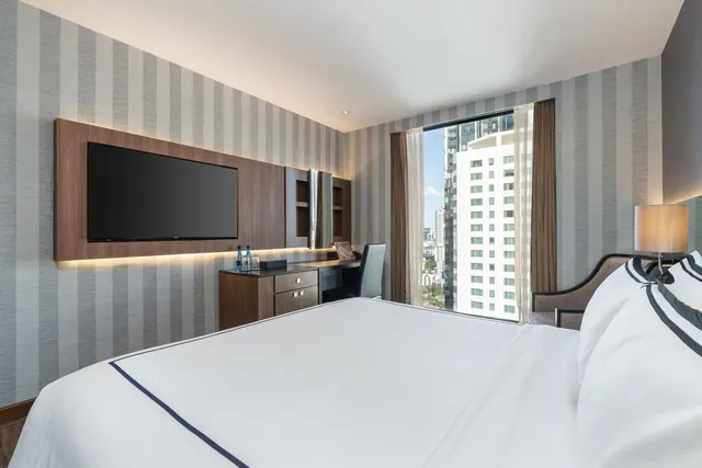 A bedroom with a made bed with navy-piped white linen, a wall-mounted television, a desk with a chair, and a window overlooking high-rise buildings.