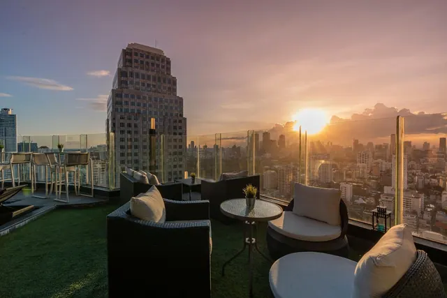A rooftop terrace with wicker armchairs, a small glass table and glass railings overlooks a city skyline at sunset.