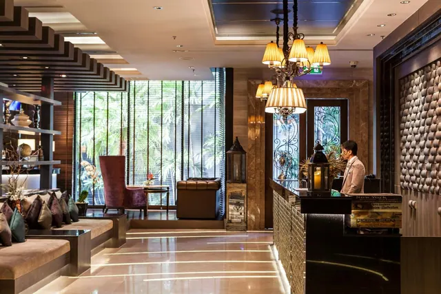 A hotel lobby with a reception desk, a hanging chandelier, lounge seating and tall windows looking onto greenery, with one staff member behind the desk.