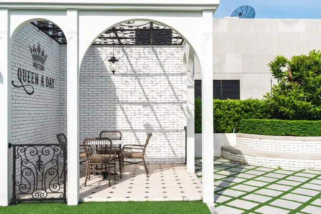 A white arched outdoor seating area holds a small table and wicker chairs, with a painted 'Queen A Day' sign on the brick wall, a green lawn and a paved path beside it.