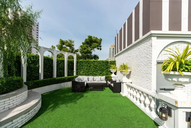 A furnished rooftop terrace with artificial turf, a grey outdoor sofa set, white pergola arches, clipped hedges and a white brick wall with a balustrade, with a tall building visible in the background.