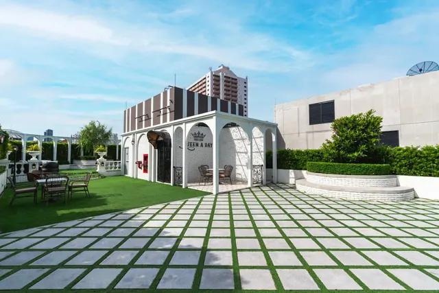 A rooftop terrace with a grass and paved grid floor, a white arched pavilion with a JEEN A DAY sign, outdoor chairs and tables, and a high-rise building in the background.
