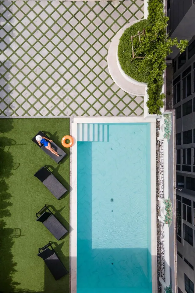 Overhead view of a rectangular pool with dark sun loungers on green turf, one guest lying on a lounger beside an orange ring float, and a lattice-patterned paved deck.