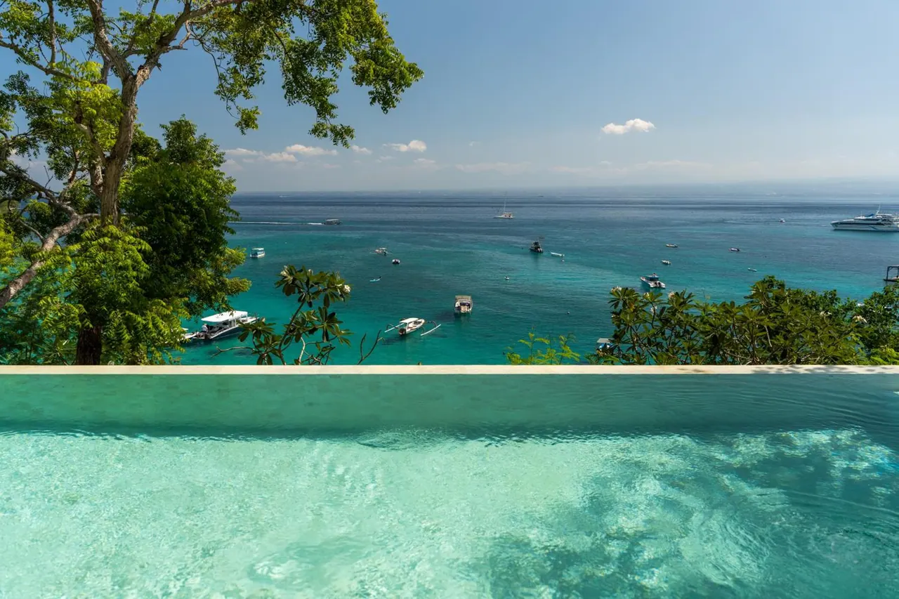 An infinity-edge pool overlooks a bay where several boats are anchored, with trees framing the left side of the view.