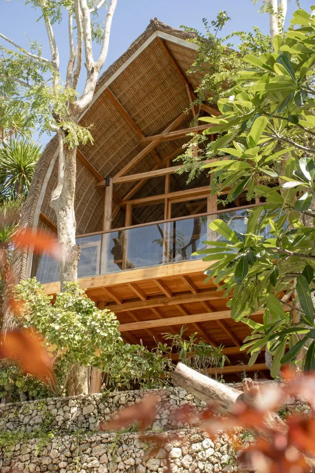 Exterior of a bamboo-roofed building with a timber upper balcony and glass railing, framed by trees and a stone retaining wall.