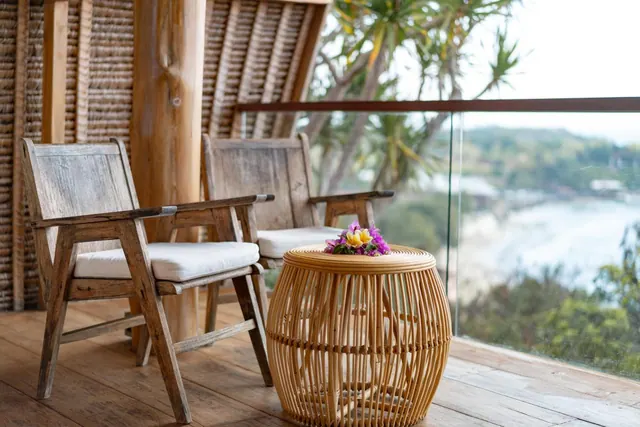 A wooden balcony with two wooden armchairs with cushions and a woven rattan side table holding flowers, overlooking trees and the sea through a glass railing.