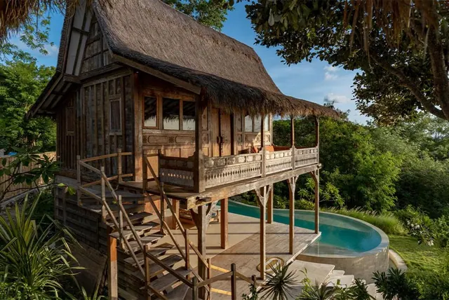 A traditional Balinese wooden house with a thatched roof, an upper balcony, exterior stairs and a curved pool to the right.
