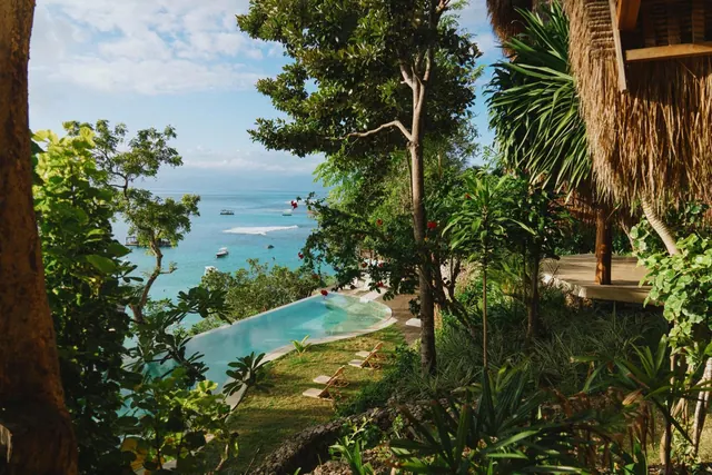An infinity-edge pool with sun loungers sits on a grassy terrace above the sea, with boats visible in the bay beyond.