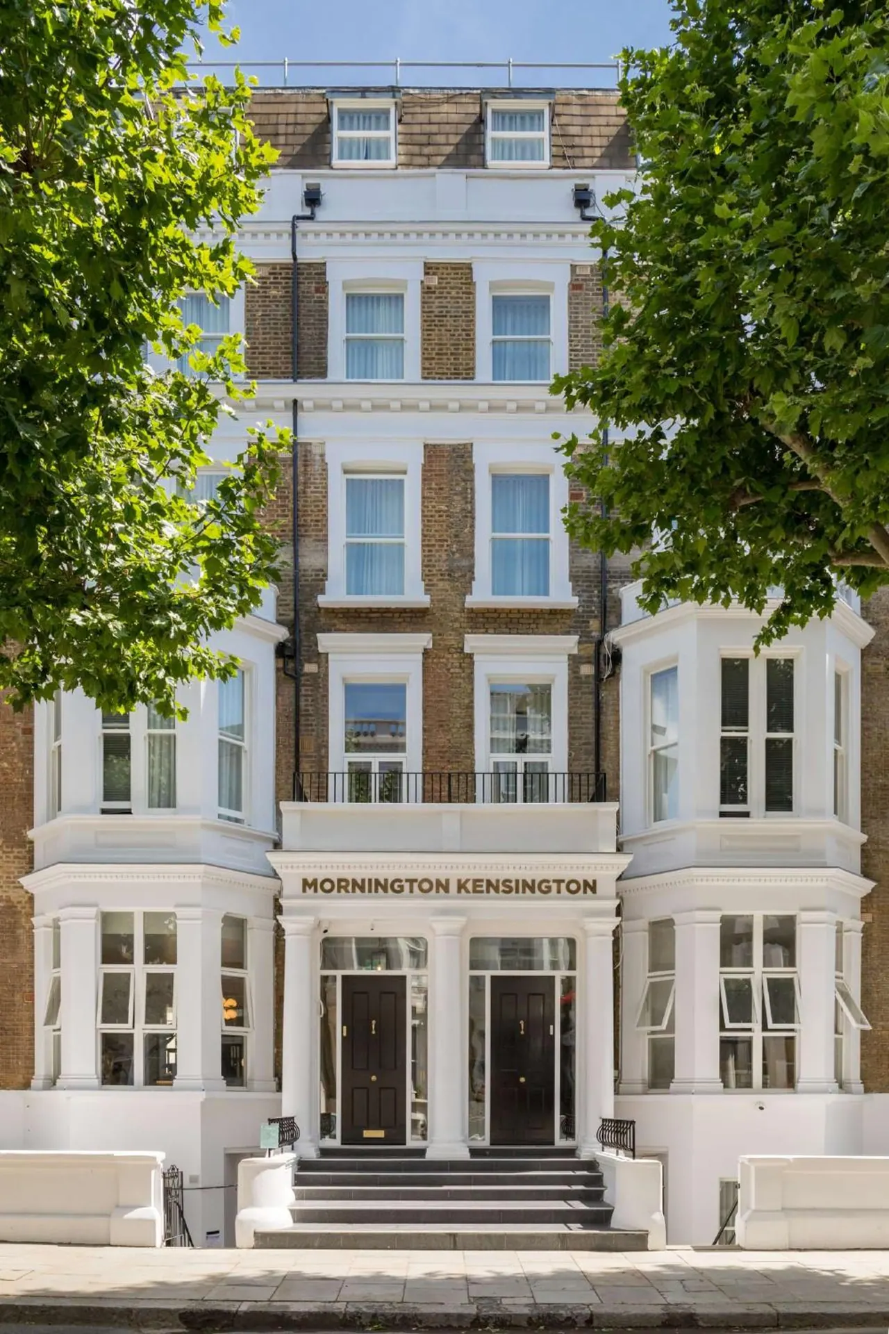 The front of the Mornington Kensington hotel, with its white-painted brick facade, columned entrance and steps, is framed by green street trees.