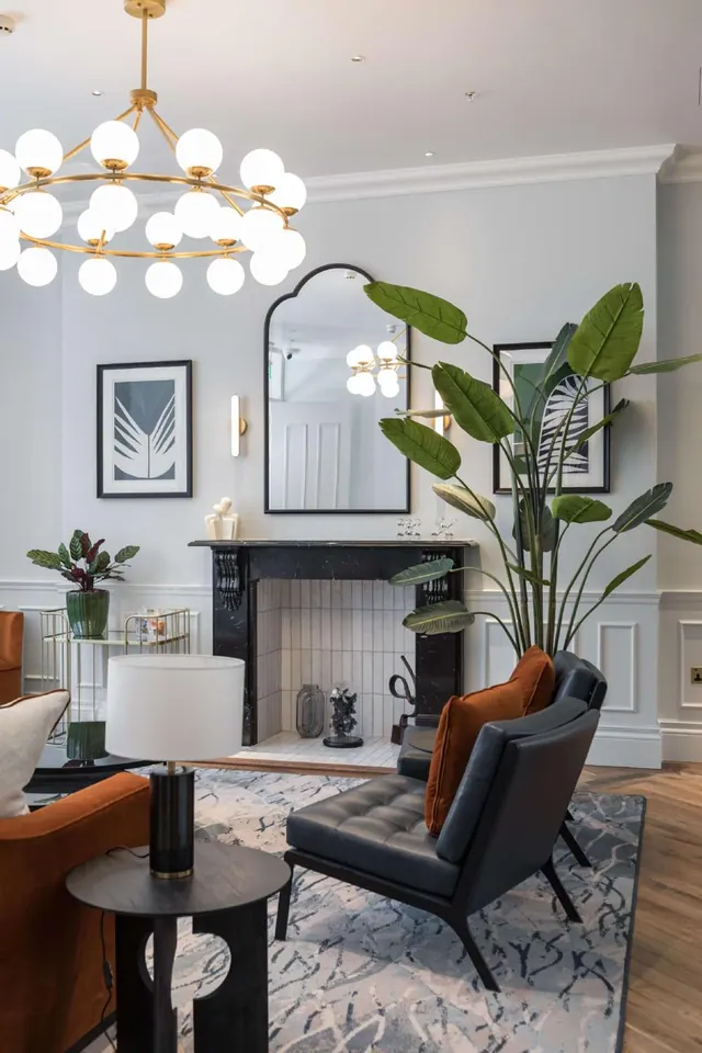 A staged lounge with two dark leather armchairs, a black marble fireplace, a large potted plant and a globe chandelier.