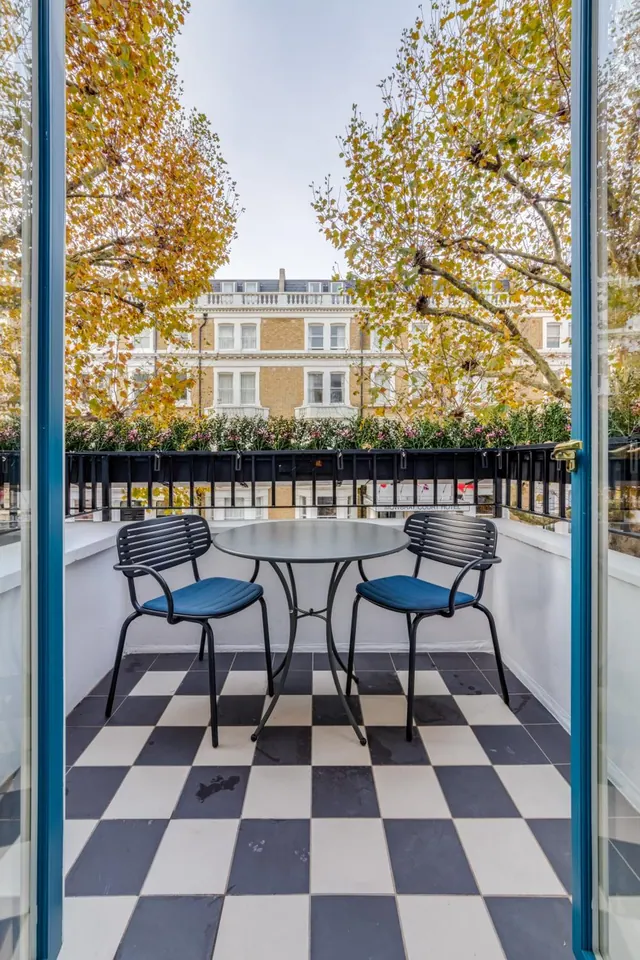 A small balcony with a black-and-white checkered tiled floor, a round table and two chairs, seen through blue-framed glass doors, with autumn trees and a building facade across the street.