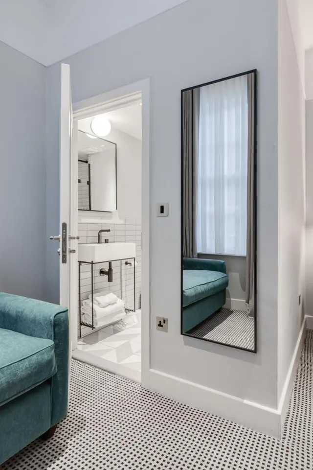 A teal armchair sits beside an open door to a white-tiled bathroom, with a tall wall mirror reflecting a window with sheer curtains.
