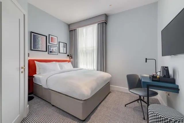 A hotel bedroom with a double bed, a wall-mounted television, and a desk with a kettle and chair beside a window.