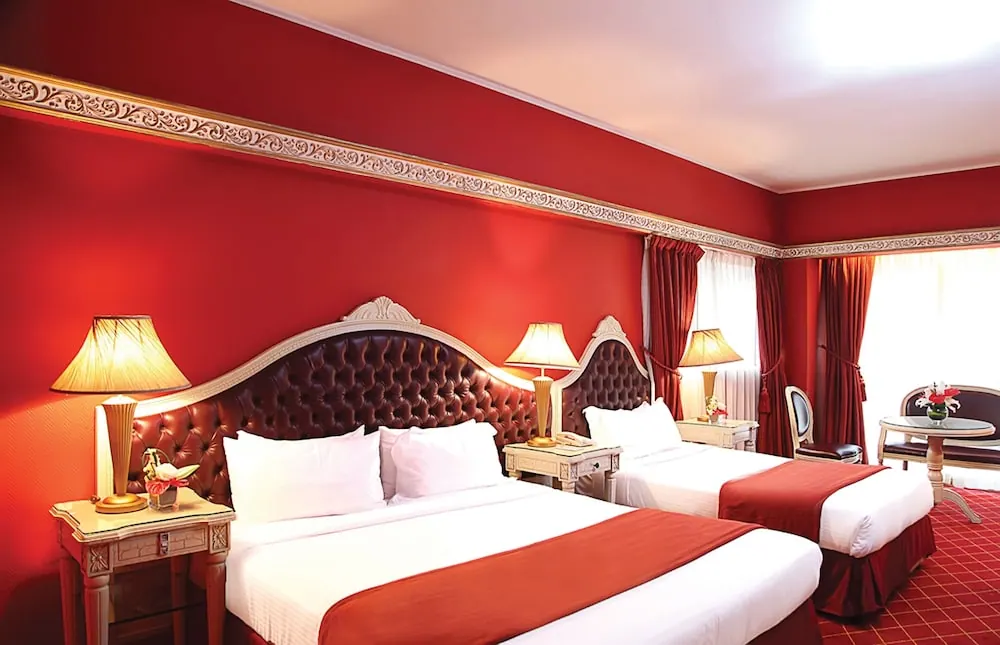 A bedroom with red walls and two double beds with tufted leather headboards, bedside lamps, curtains, and a round table with chairs near the window.