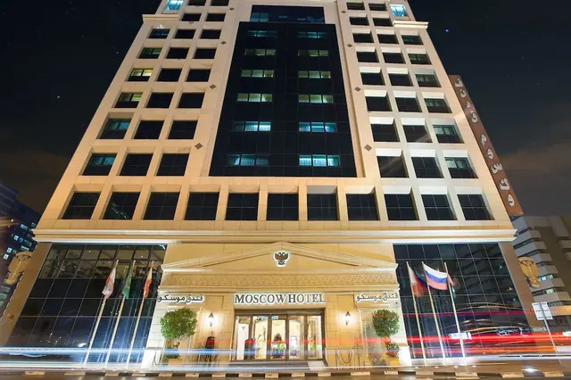 Night exterior of the Moscow Hotel building with its illuminated entrance, flags, and a vertical sign on the side of the facade.