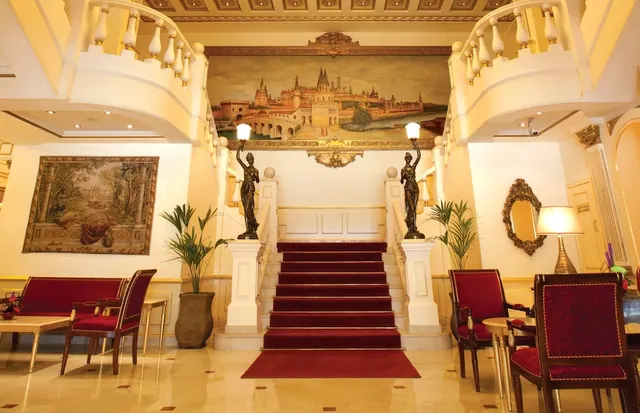 A lobby with a central staircase covered in red carpet, flanked by bronze figure statues holding lamps, a large painted scene above the stairs, a tapestry on the left wall, and red upholstered seating.