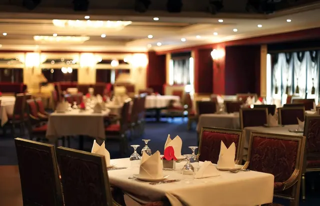 A formal dining room with white-clothed tables set with folded napkins and wine glasses, dark red upholstered chairs, chandeliers and recessed ceiling lights.