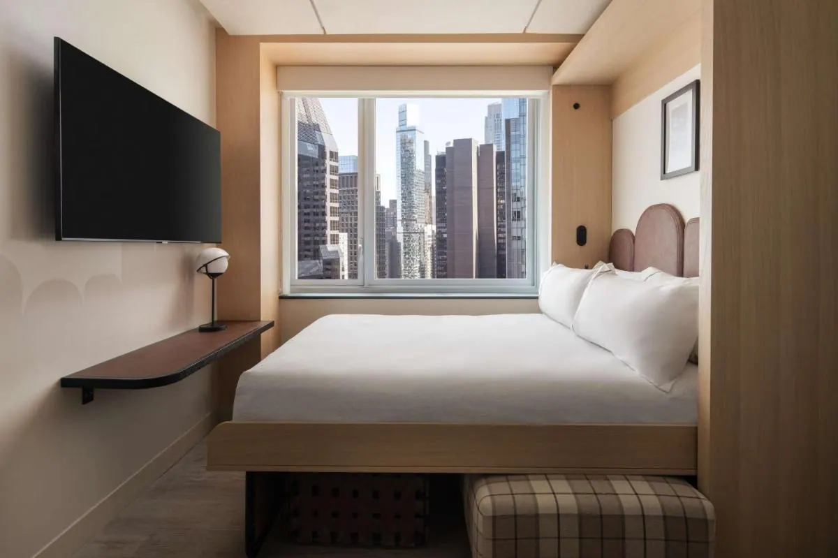 A bed with white linens sits beside a window showing high-rise buildings, with a wall-mounted television on the left.