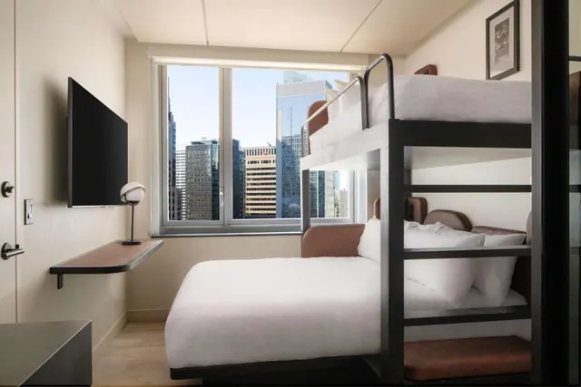 A small hotel bedroom with a bunk bed above a double bed, a wall-mounted television, and a window looking out over city skyscrapers.