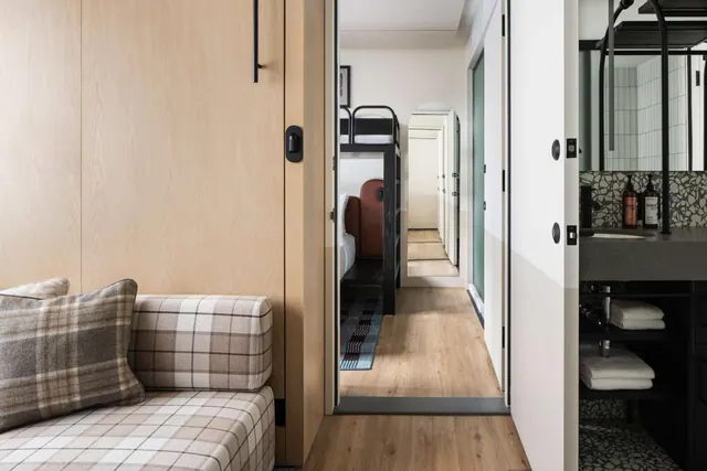 A plaid upholstered sofa sits beside a wood-panelled wall, with a view down a hallway to bunk beds and a bathroom vanity with folded towels on the right.