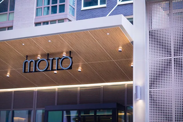 The hotel entrance canopy shows the Motto name sign on a wood-panelled ceiling with warm downlights, beside a perforated metal wall.