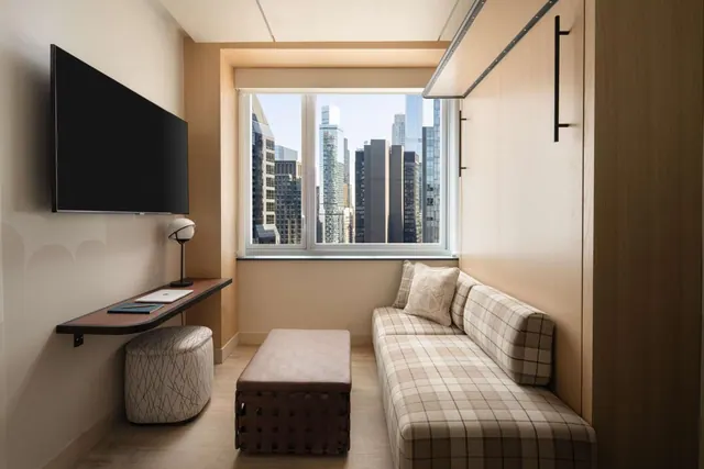 A small seating area with a wall-mounted television, a wall desk with a lamp and laptop, a plaid sofa, and a window overlooking city skyscrapers.
