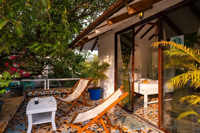 A furnished private terrace with loungers overlooks tropical greenery and a pool, with the bedroom visible through glass doors.