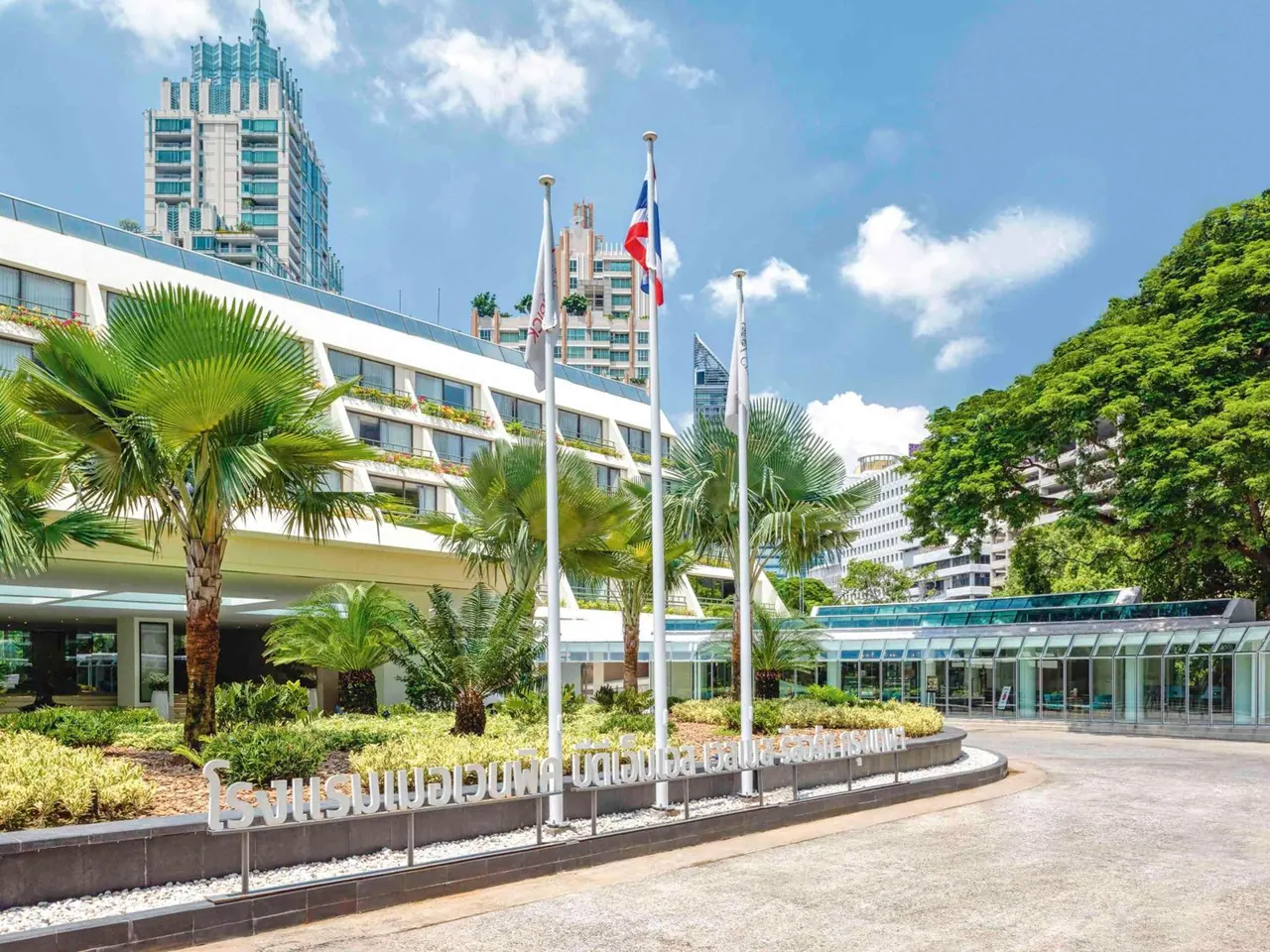 The hotel entrance has two flagpoles, palm trees, a metal sign with Thai lettering, a glass-walled pavilion and a curved driveway under a blue sky with tall buildings behind.