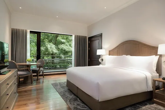 A bedroom with a king bed, a wooden dresser with a television, a round table with two chairs, and a glass door opening onto a balcony overlooking green trees.