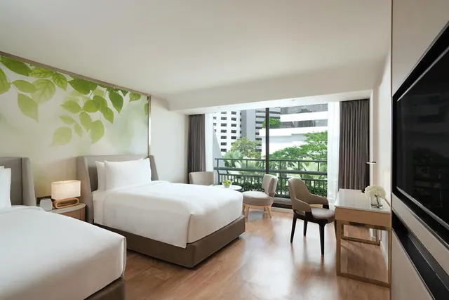 A hotel bedroom with two single beds, a leaf mural on the wall, armchairs and a desk by a large window that looks onto a balcony and green trees.