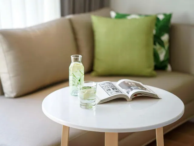 A beige sofa with green cushions and a white round side table holding a carafe of cucumber water, a glass and an open magazine.