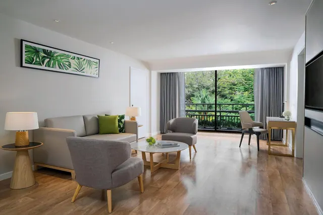A living area with a grey sofa, grey armchairs, a round coffee table and a wall-mounted television, with floor-to-ceiling glass doors opening onto a balcony with trees beyond.