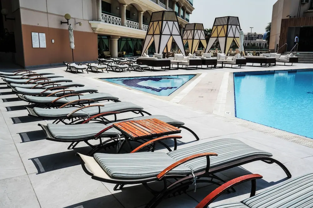 Rows of striped sun loungers line the pool deck beside a large blue pool and a small tiled pool, with cabana seating and a hotel building behind.