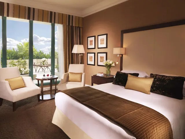 A hotel bedroom with a king bed, two armchairs and a round side table, with floor-to-ceiling windows opening onto a balcony with tropical greenery beyond.