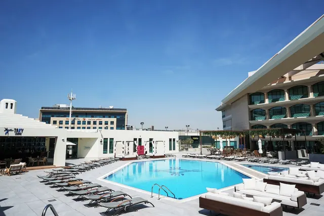 An outdoor pool with rows of sun loungers along its left edge, white cushioned daybeds in the foreground, and a multi-storey hotel building on the right under a clear blue sky.