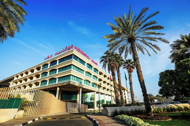 The exterior of a multi-storey Movenpick Grand hotel building with its name on the facade, flanked by tall palm trees under a clear blue sky.