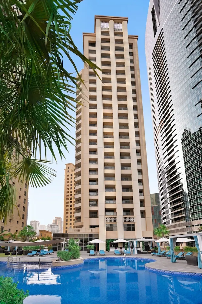 A large outdoor pool with blue loungers and white umbrellas sits in front of a tall beige residential tower and a glass skyscraper.